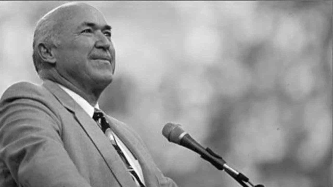 Pastor Chuck Smith in a light suit, looking up and slightly smiling, speaking at an outdoor microphone. Black and white.