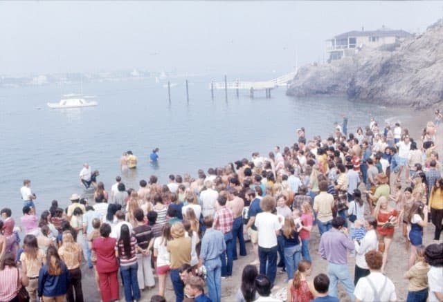 A crowd of young people on a beach watching baptisms in the shallow water, a rocky point and a house behind them.