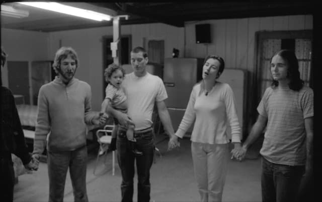 Five young adults, one holding a small child, standing in a circle holding hands to pray. Black and white.