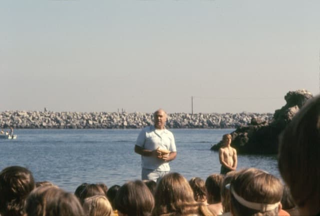 Pastor Chuck Smith at the water's edge, a book in his hands, speaking to a crowd on the sand, a breakwater behind him.